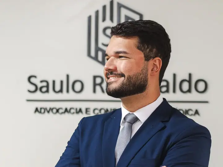 Saulo Romualdo diante da placa do escritório, de braços cruzados